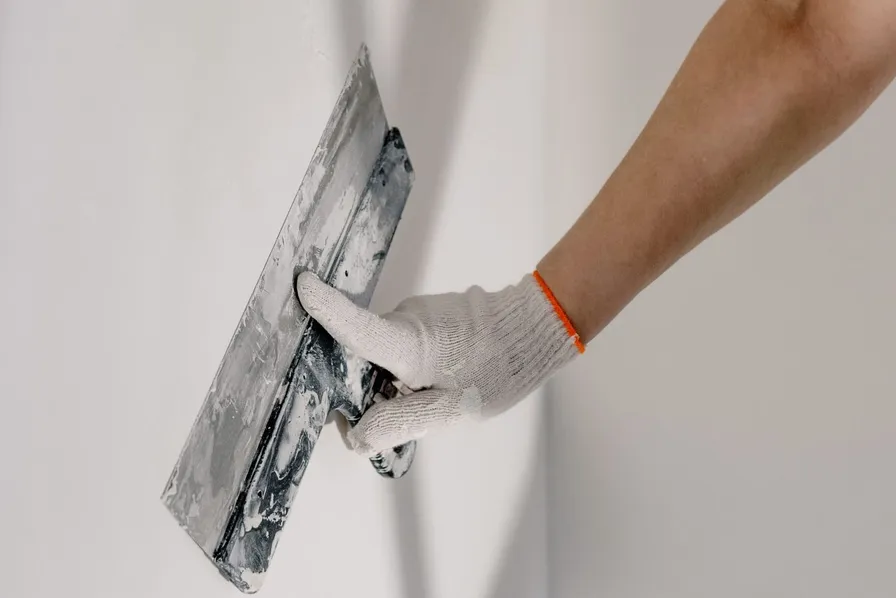 A wide taping knife in a gloved hand gliding over a smooth finished drywall wall