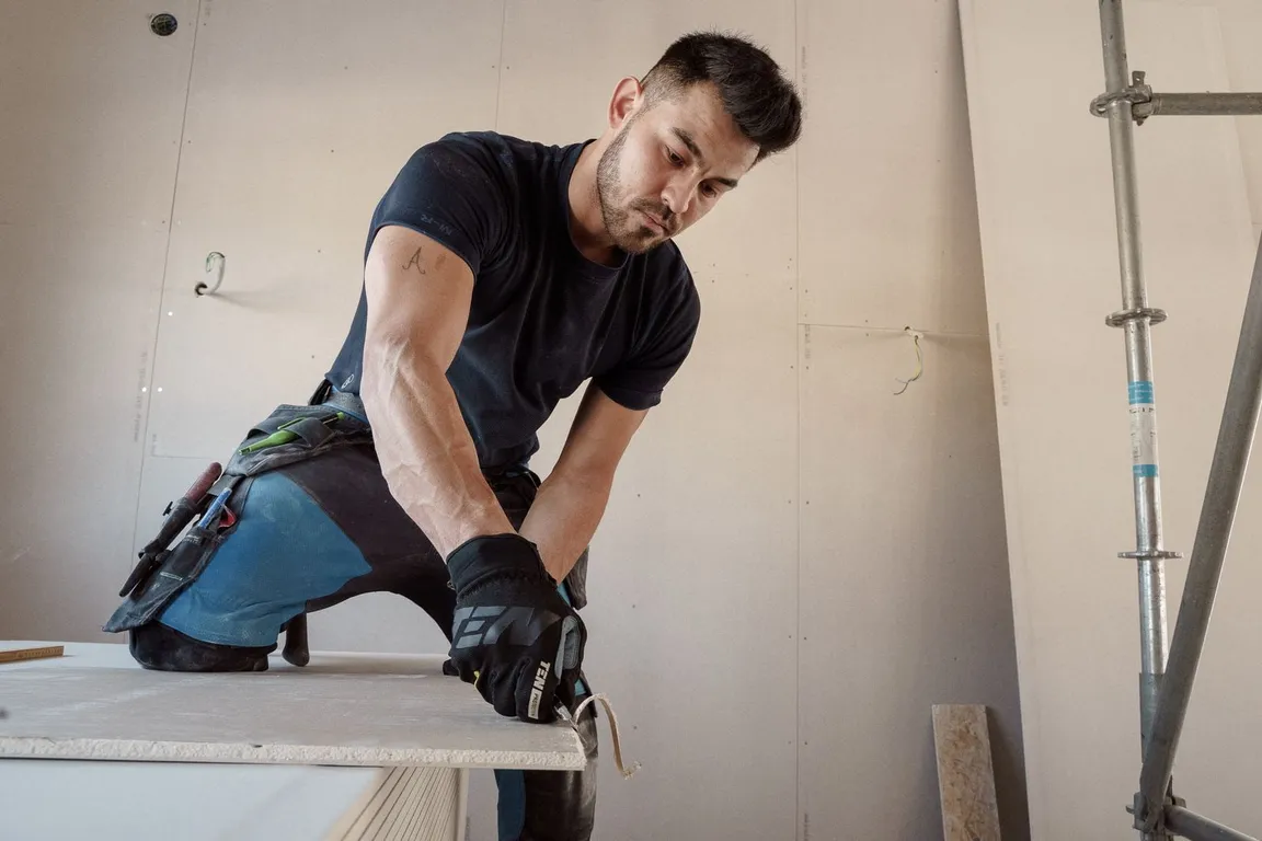A drywall contractor kneeling on a stack of sheets, cutting a board to size in a freshly boarded room