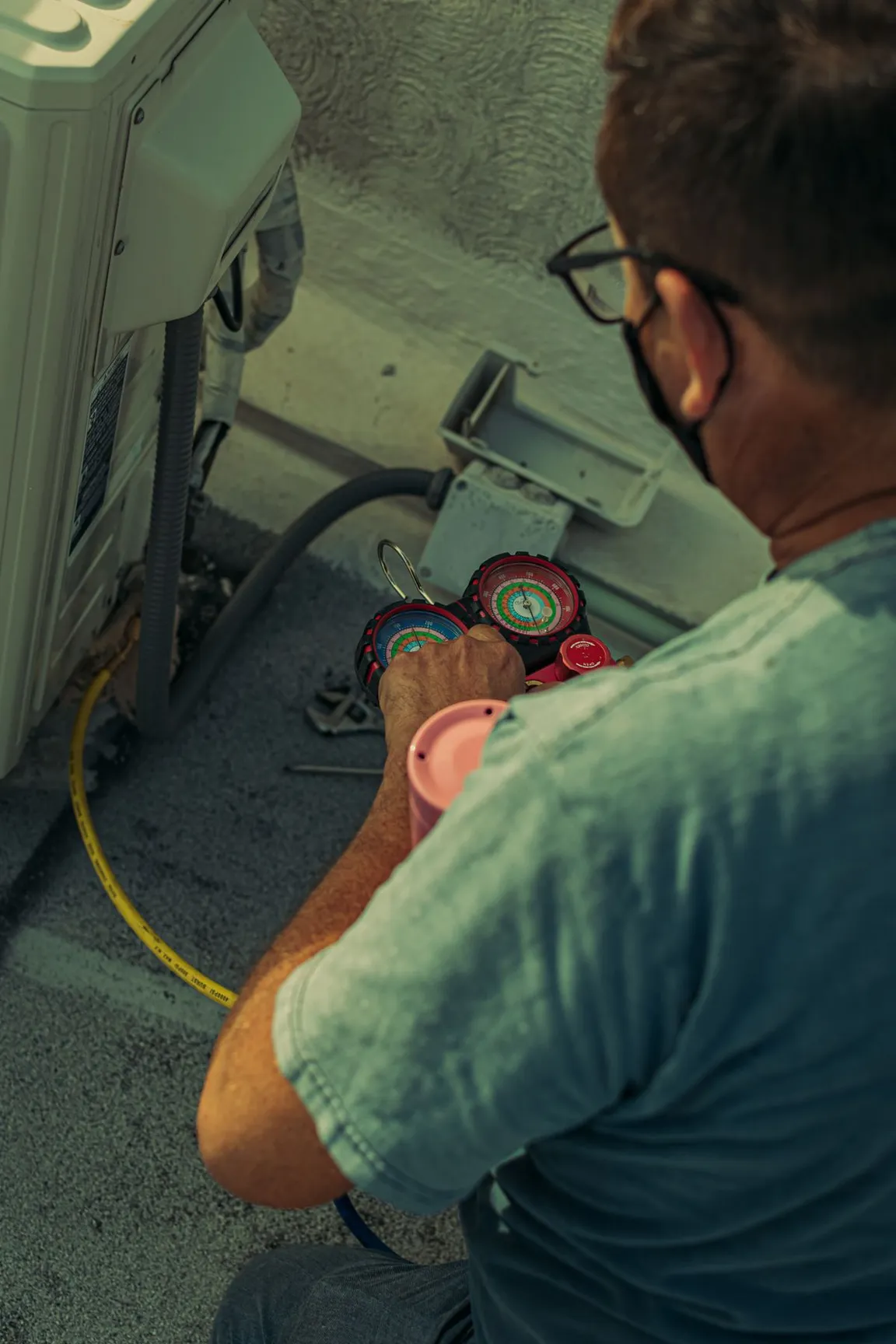 An HVAC technician kneeling at a condenser unit reading manifold gauges