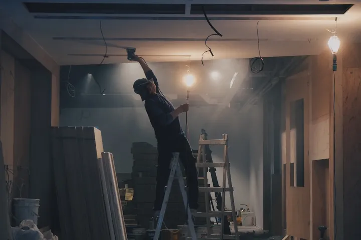 A tradesman up a ladder working under jobsite lights