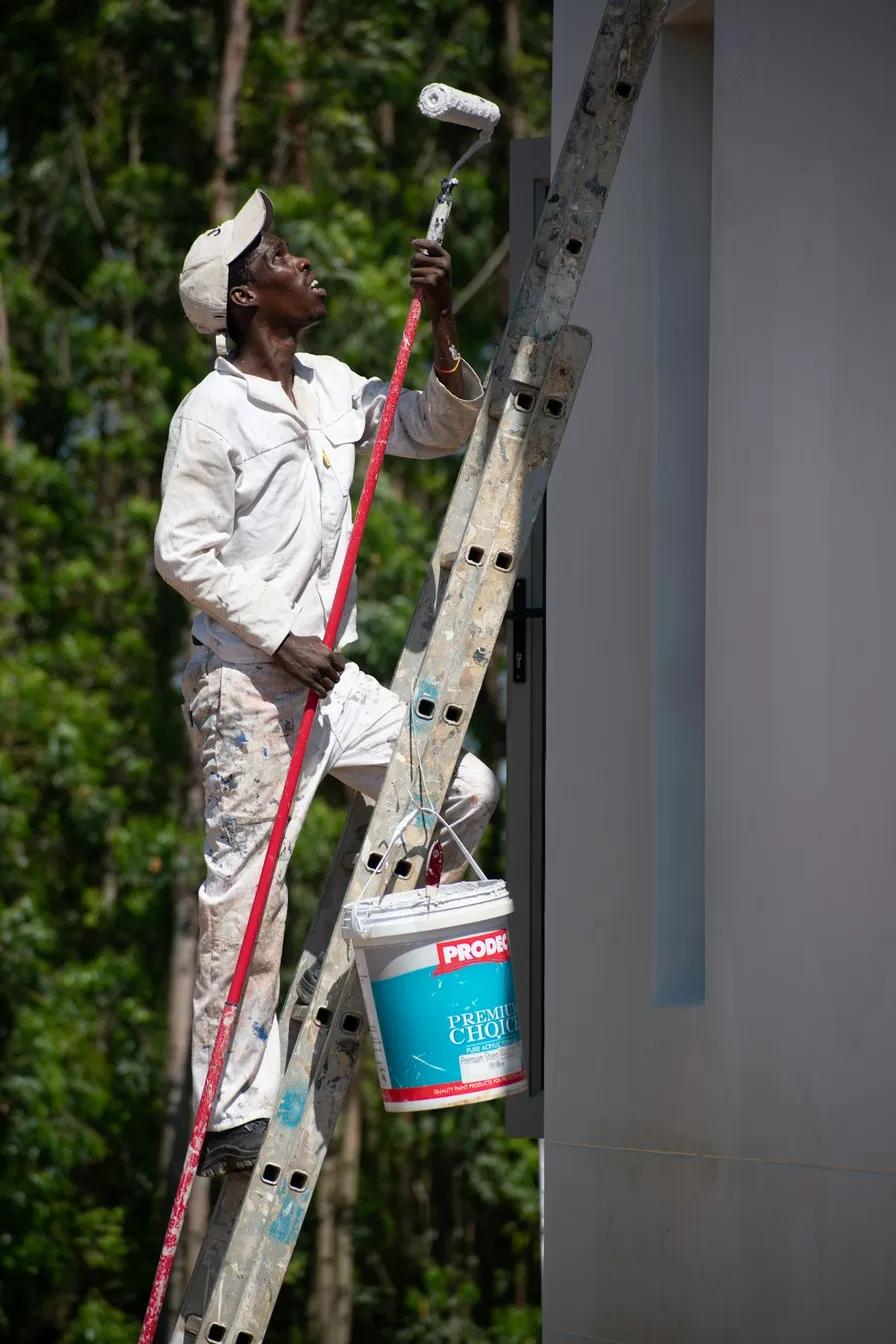 An exterior painter working up an extension ladder, roller pole in hand and paint bucket hooked below